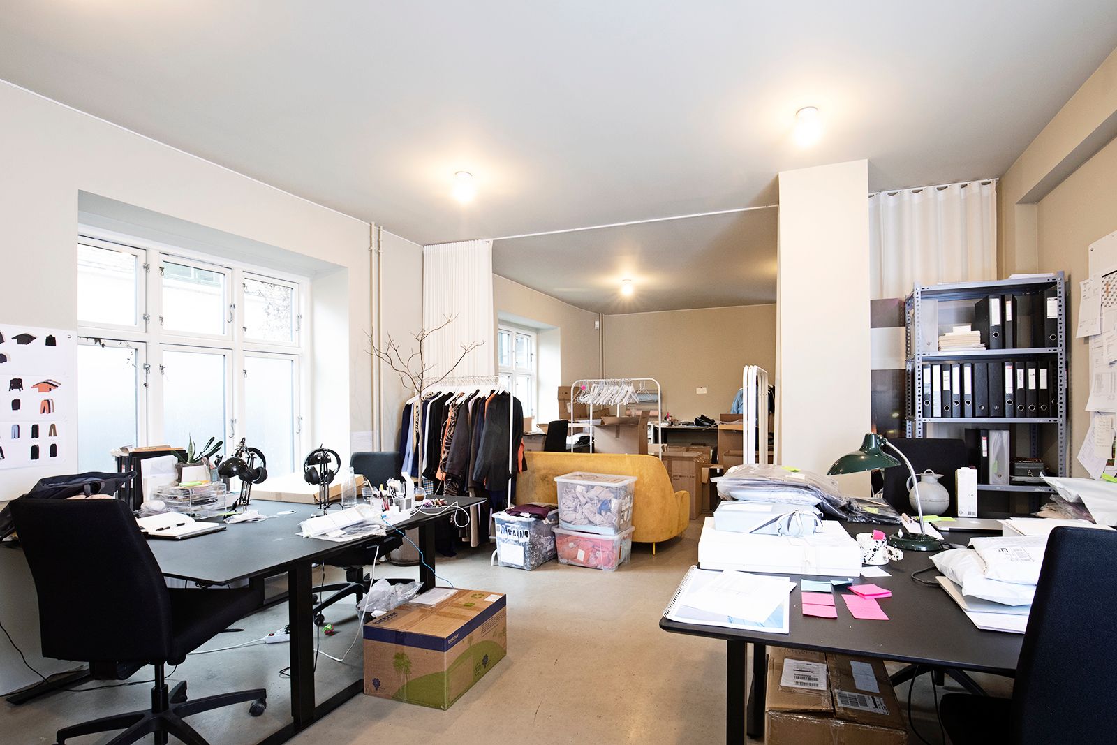 Kontor med to bord, en gul sofa og en høj, hvid hylde med mapper. Lyse lofter og gulv samt en åben gardin og vinduer med lys.