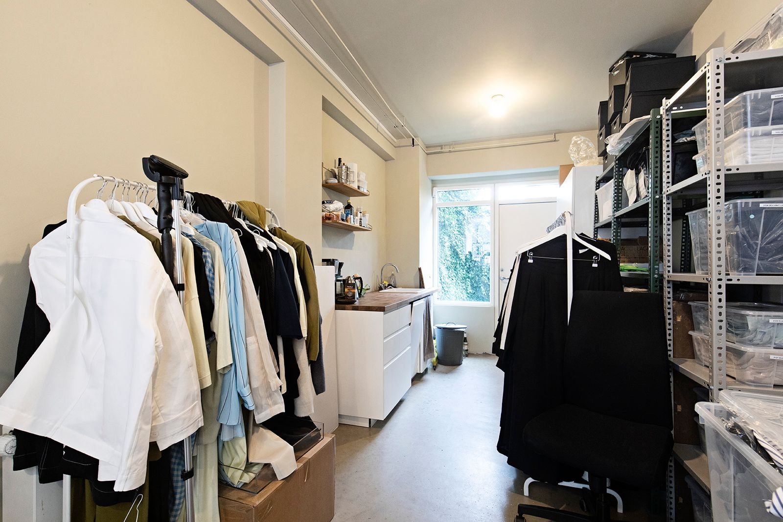 En lille, velorganiseret garderobe med hængehylder og skraldspand samt et køkken med lyse møbler og en stor åbning mod