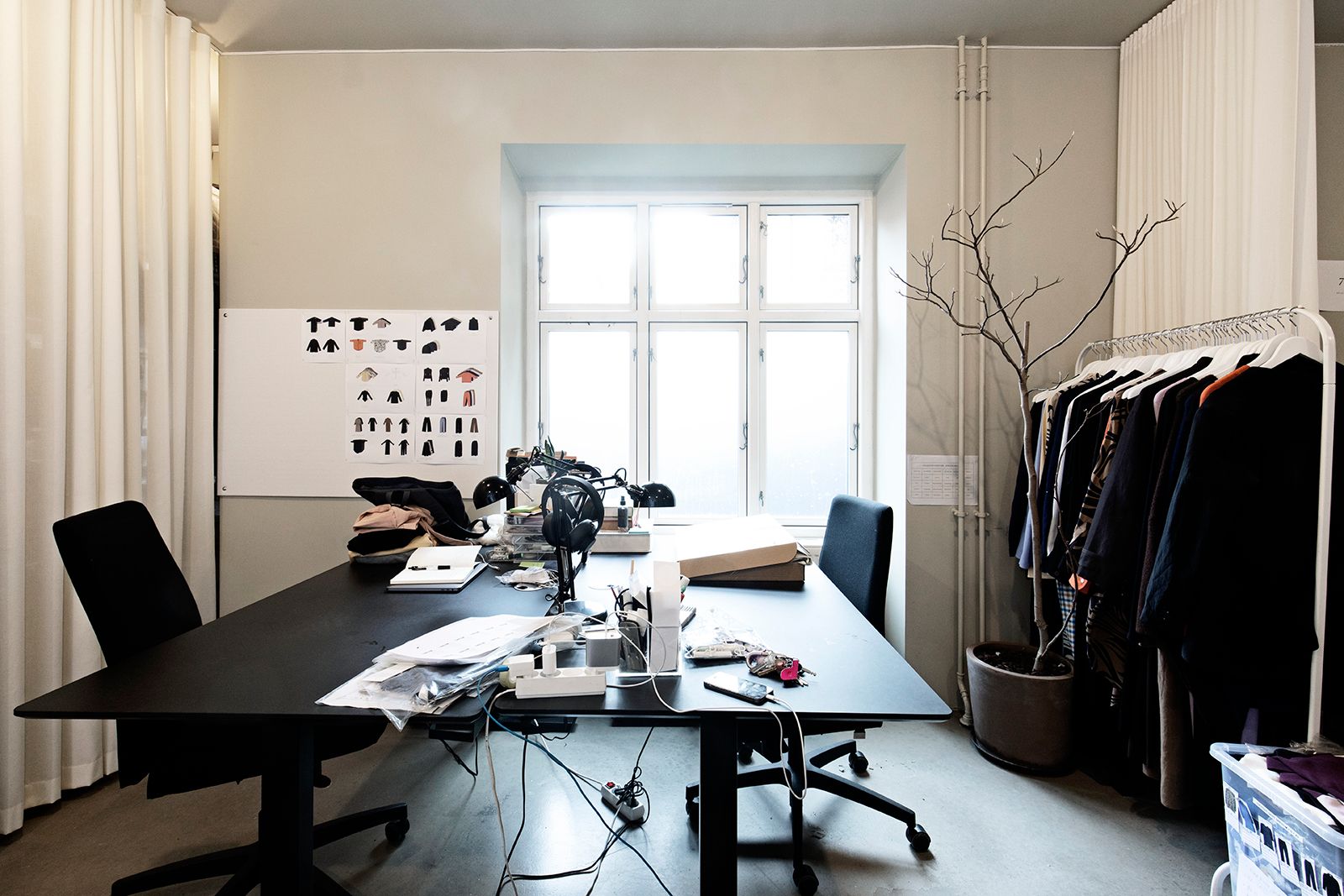 Et arbejdsrum med to skriveborde og stole og et hængeskab med tøj samt et whiteboard med tøjbilleder