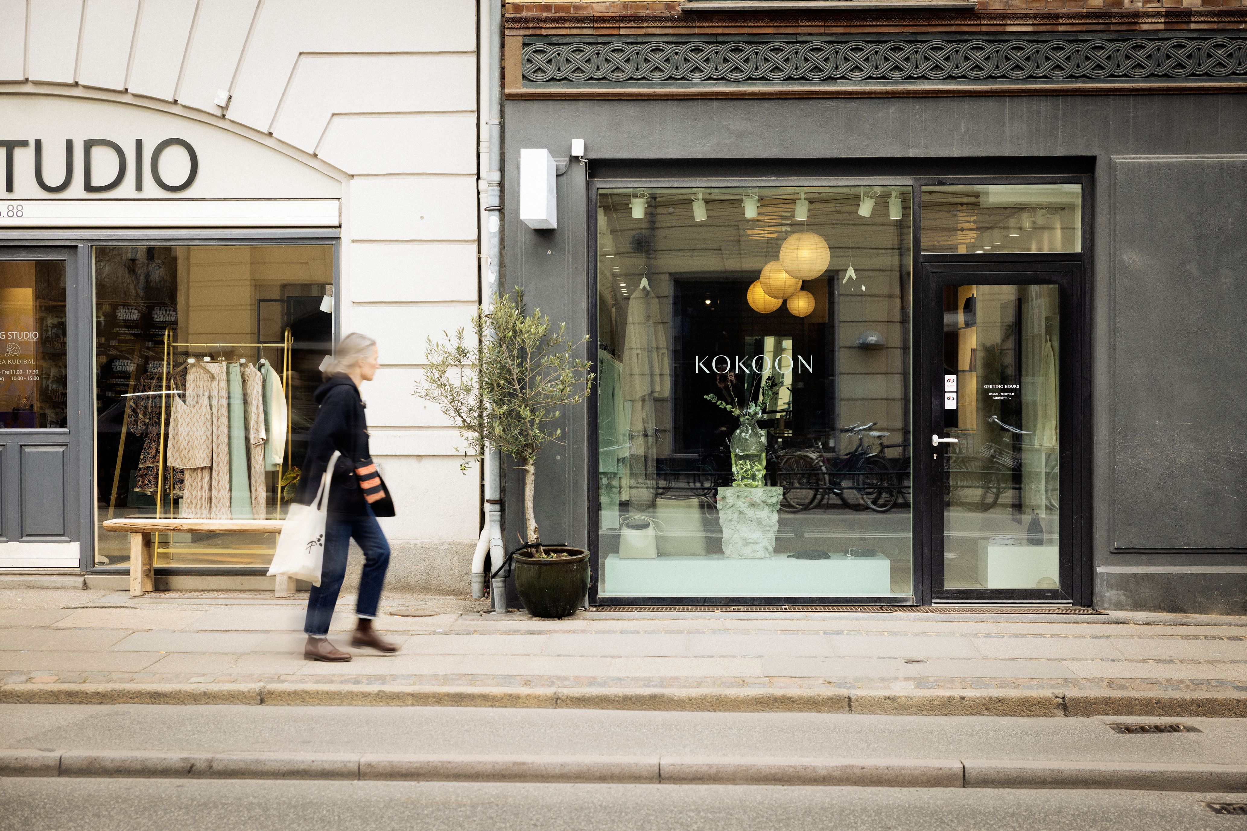 Personer går forbi en butik med navnet Kokoon og et studie på en gågade med træplanter i potter og køretøjer i udsigten