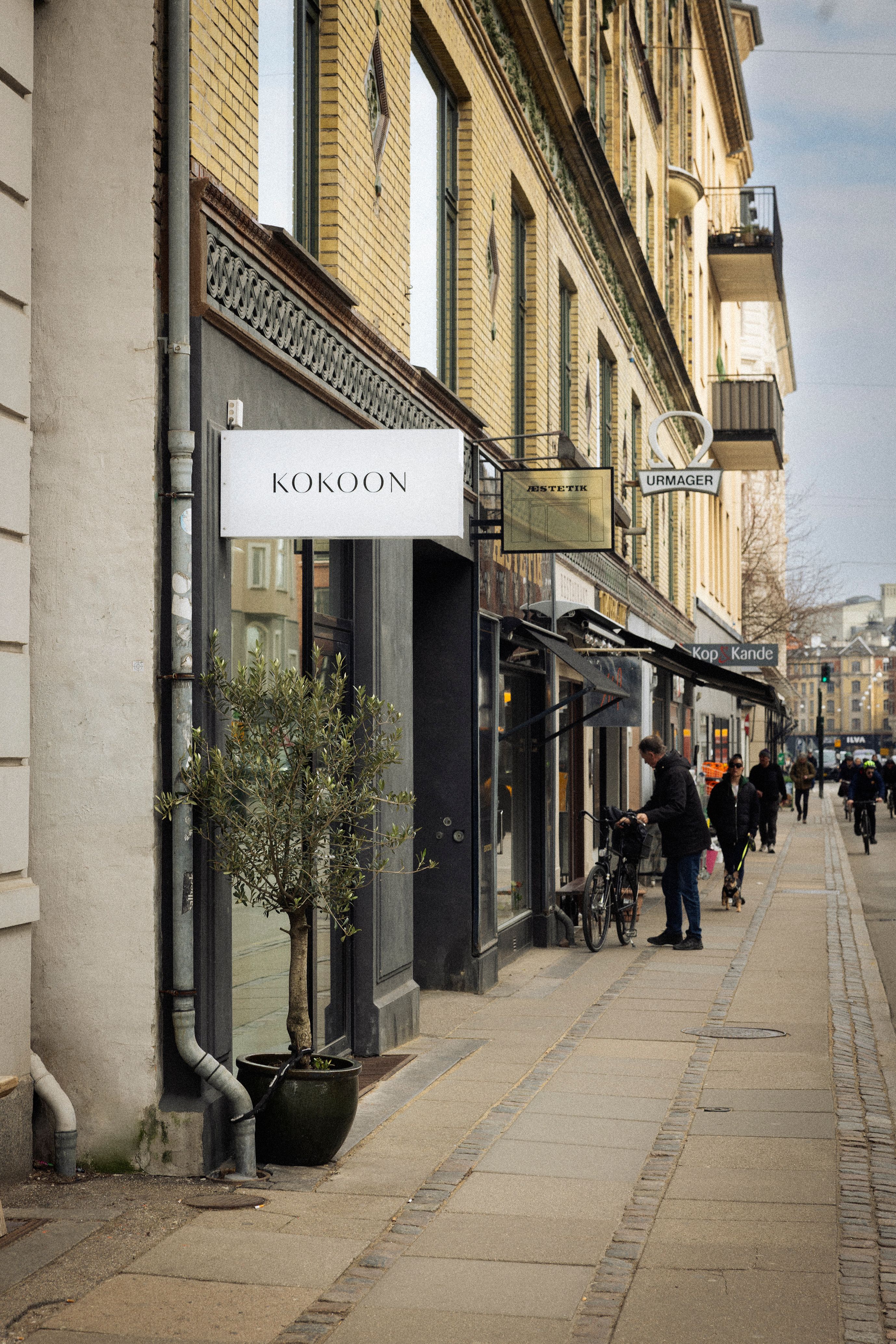 Butik med navnet Kokoon placeret på gågaden, hvor folk passerer med cykler og hunde