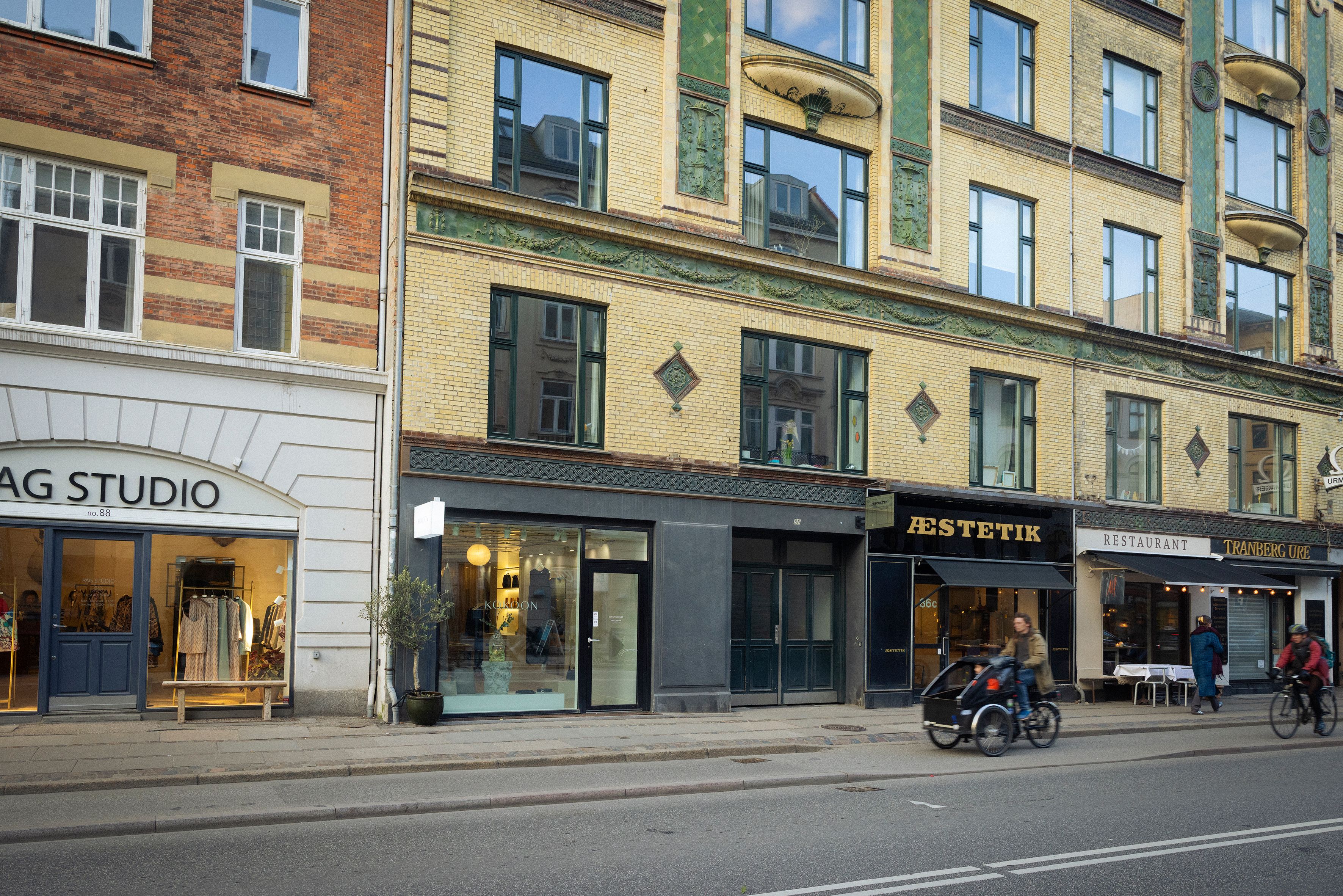 Mennesker kører med cykel og vogn, og en person cykler udenfor. Der er flere forretninger med butiksruder og tekster