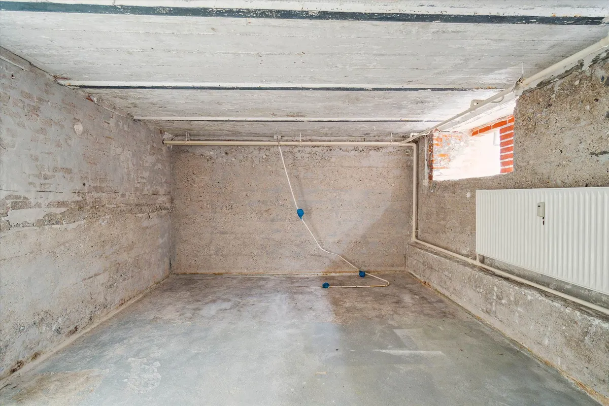 En tom garage med hvide rører og kabel, cementvæg og gulv, hvid radiator og et lille vindue med rød kant.