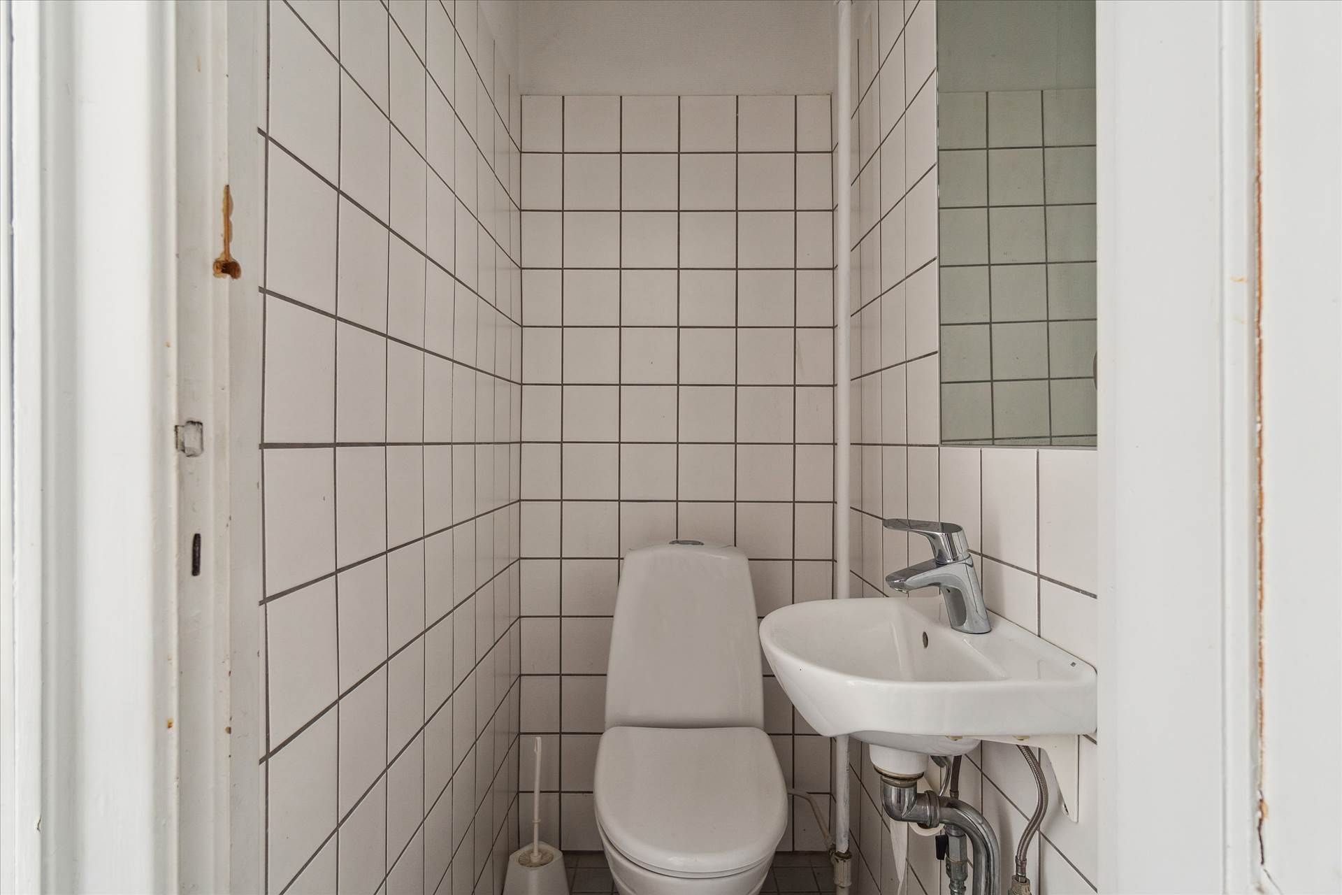 Et lille, hvidt badeværelse med toilet, håndvask og spejl samt et badeværelsesbørste på gulvet