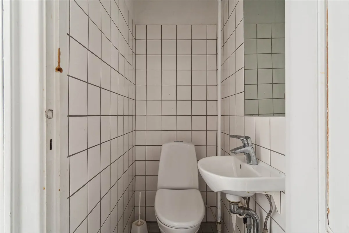 Et lille, hvidt badeværelse med toilet, håndvask og spejl samt et badeværelsesbørste på gulvet