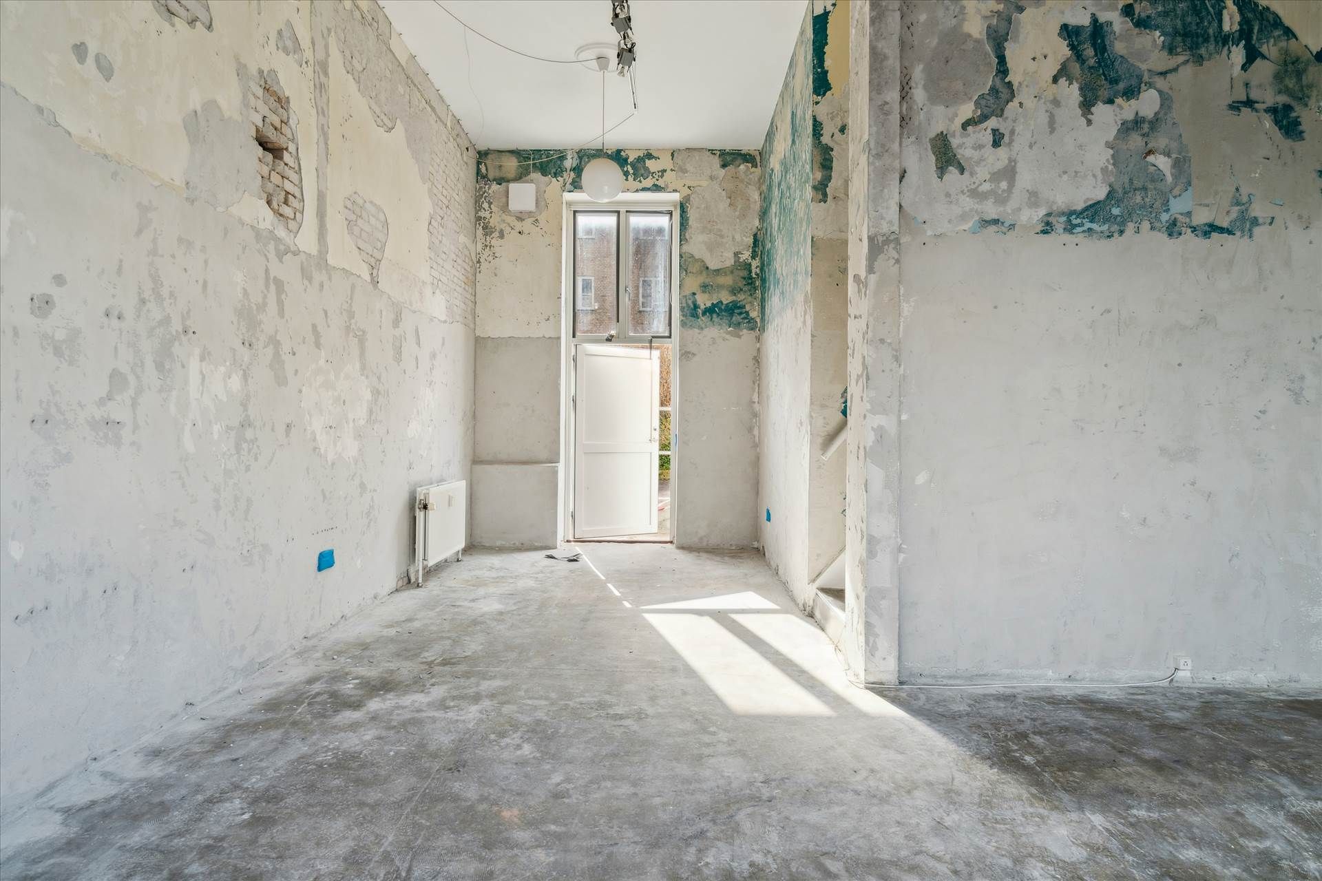 Et tomt værelse med revne vægge og hul i væggen og åben dør, der fører udenfor og til trapper, samt solstråler på gulvet