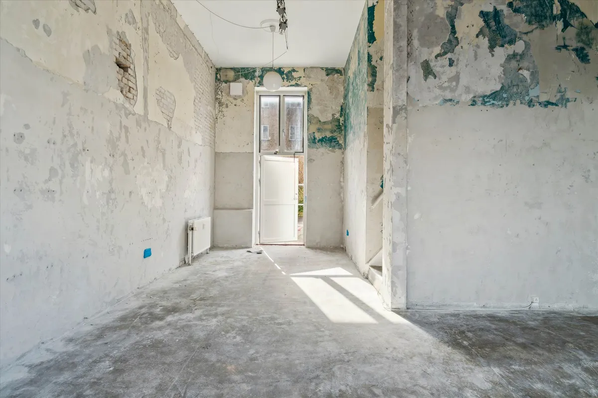 Et tomt værelse med revne vægge og hul i væggen og åben dør, der fører udenfor og til trapper, samt solstråler på gulvet