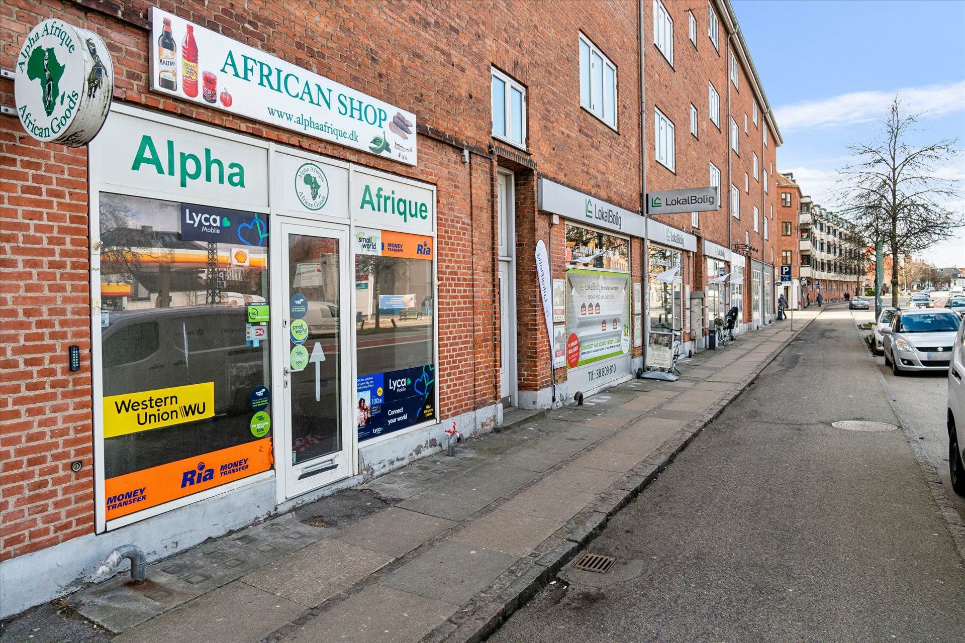 Gade med to butikker og en parkeringsplads, hvor den ene butik hedder Alpha African Shop og den anden LokalBolig, og flere