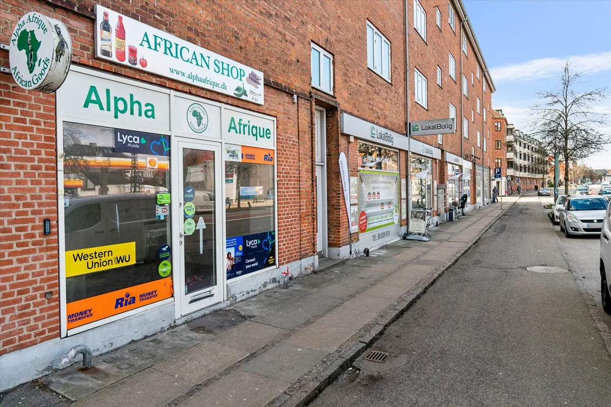 Gade med to butikker og en parkeringsplads, hvor den ene butik hedder Alpha African Shop og den anden LokalBolig, og flere