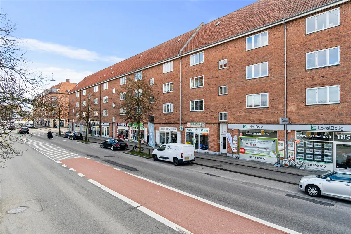 Gadebillede med biler og en bygning med butikker og Lokalbolig skilte. I baggrunden er der en tankstation og flere biler