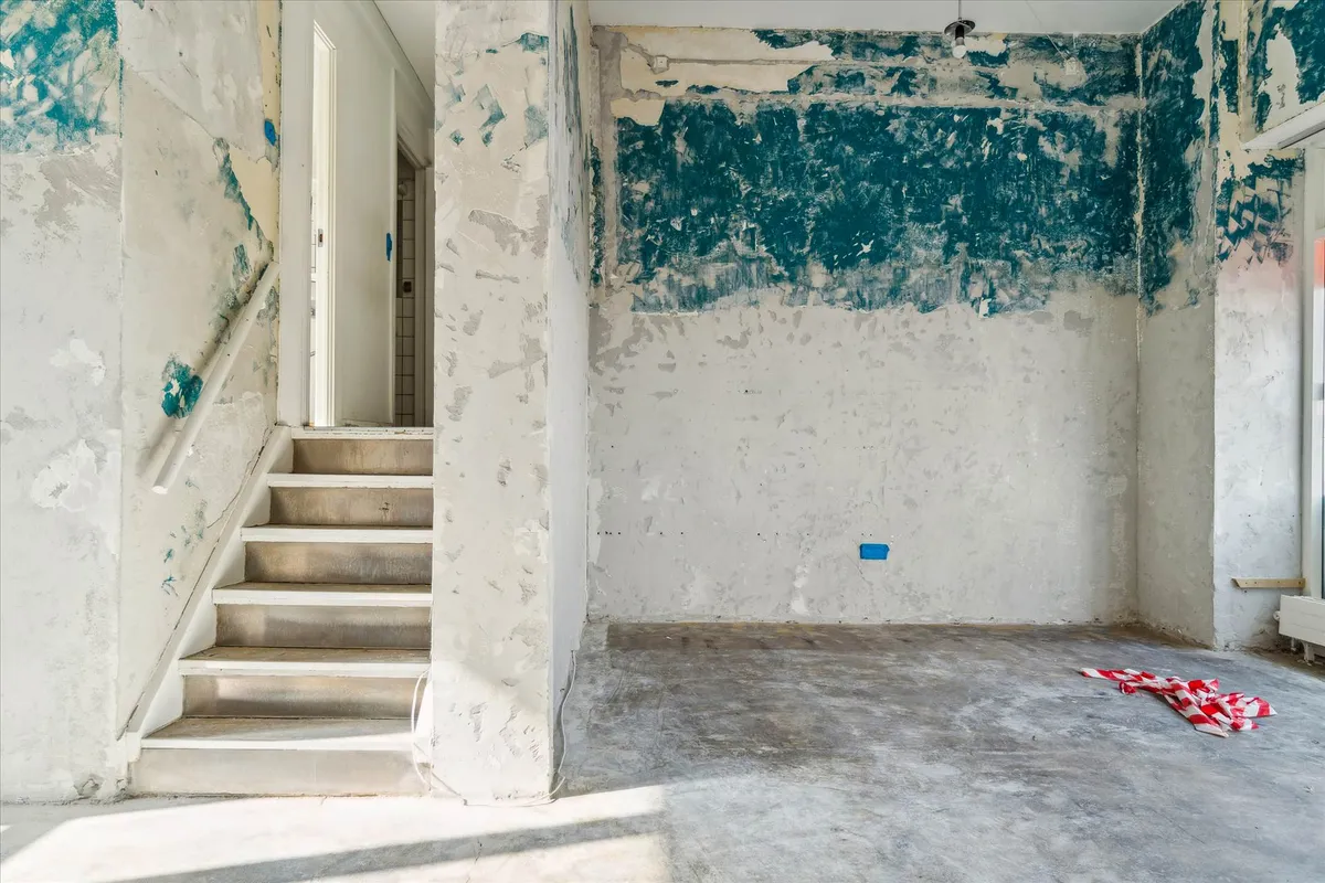 En ubeboet stue med et forfaldent loft og vægge. Der er et trin til en trappe med hvidt gelænde. Væggen er slettet med