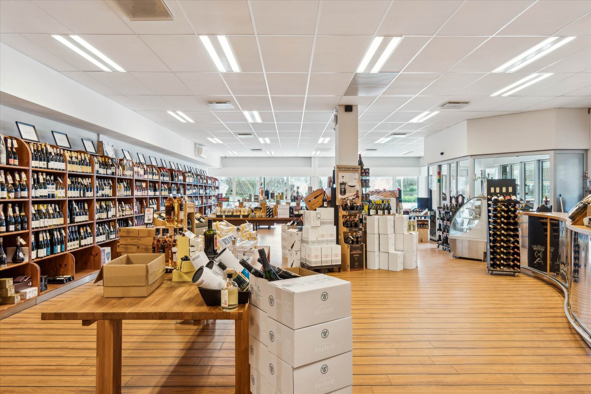 Indenfor en vinforretning er der flere høje hylder med flasker og bokse med vin og andre alkoholiske drikkevarer samt en