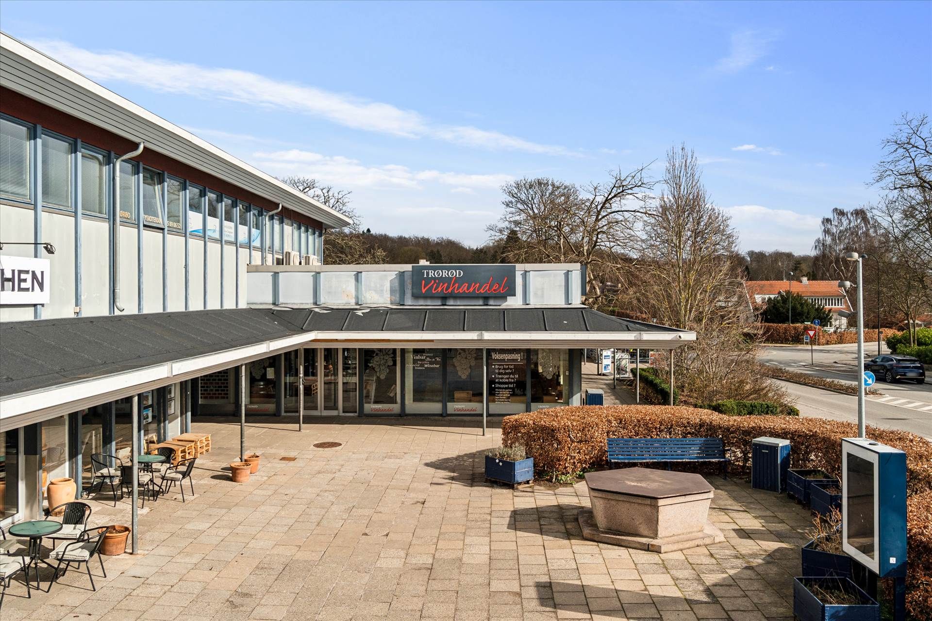 Trorod Vinhandel med udendørsborde og stole, buske og møbler samt en søjle med skilt og parkeringsplads