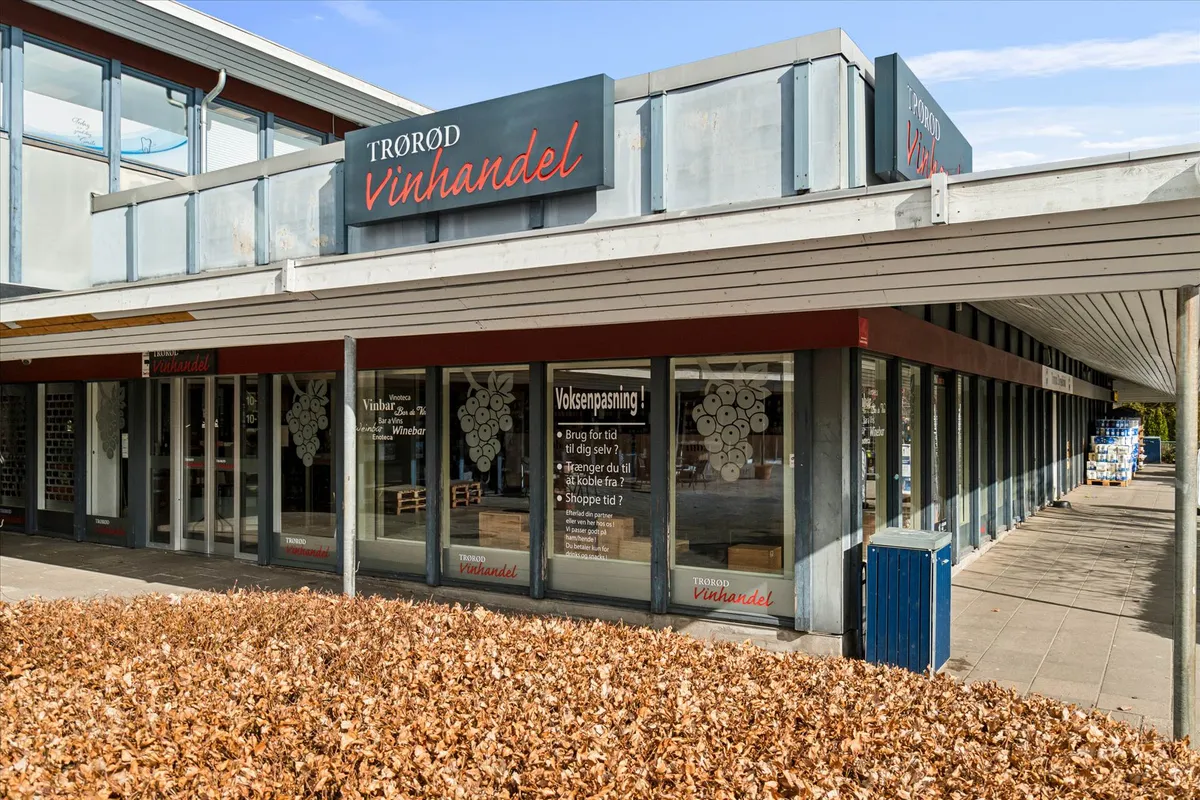 Trorod Vinhandel står i en hvid bygning med flere vinduer og røde bogstaver. Et blåt søjlesystem står foran bygningen og