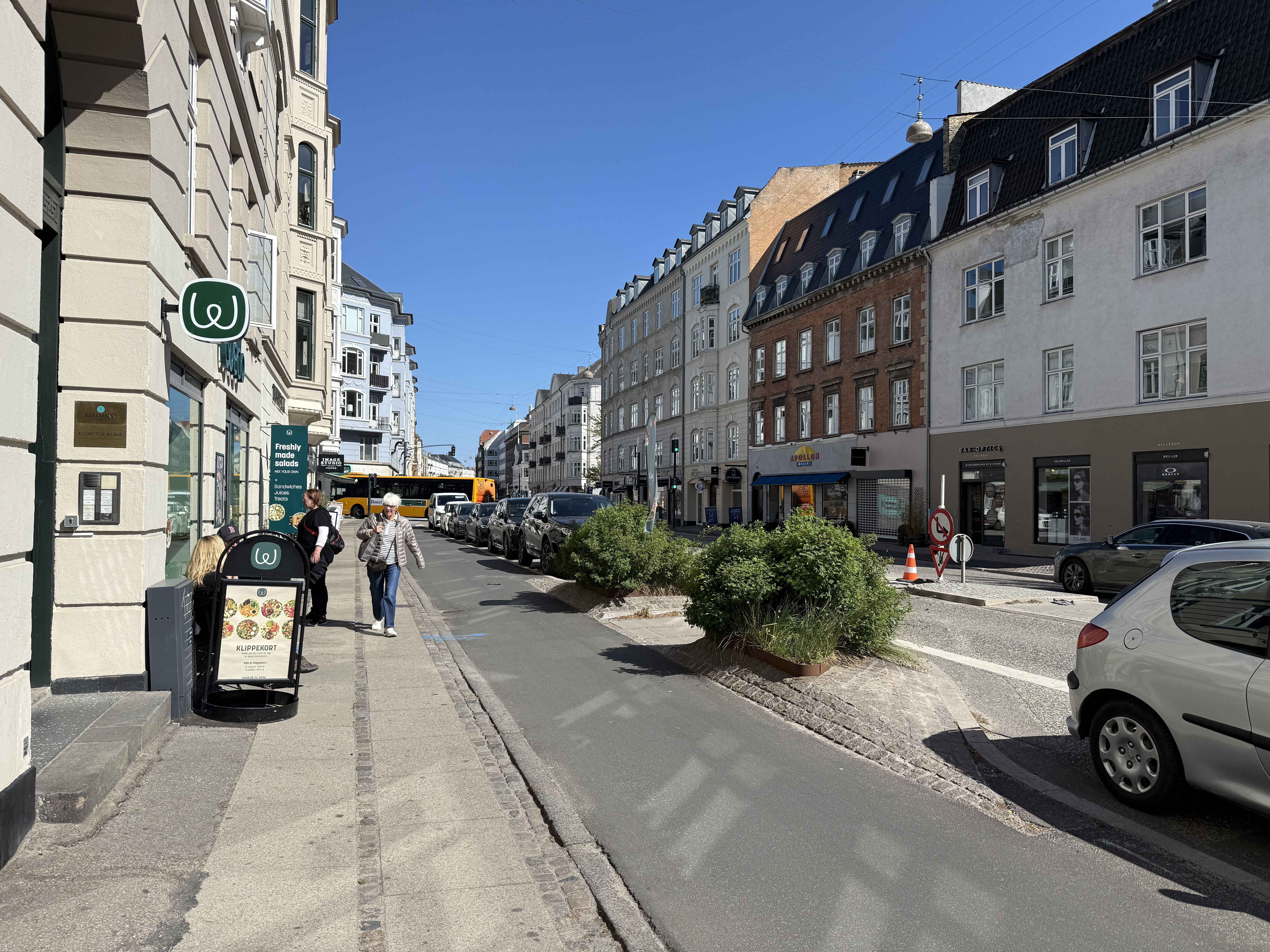 Gågade med kantsten og busser på Strandvejen. To personer går mod hinanden. Forskudte plantager og udsalgsskilte.