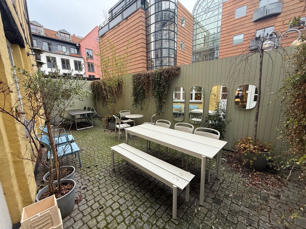 En terrasse med hvide borde og stole og plantepoter samt tre spejle i en byggegrund med huse bagved