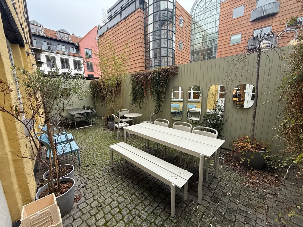En terrasse med hvide borde og stole og plantepoter samt tre spejle i en byggegrund med huse bagved