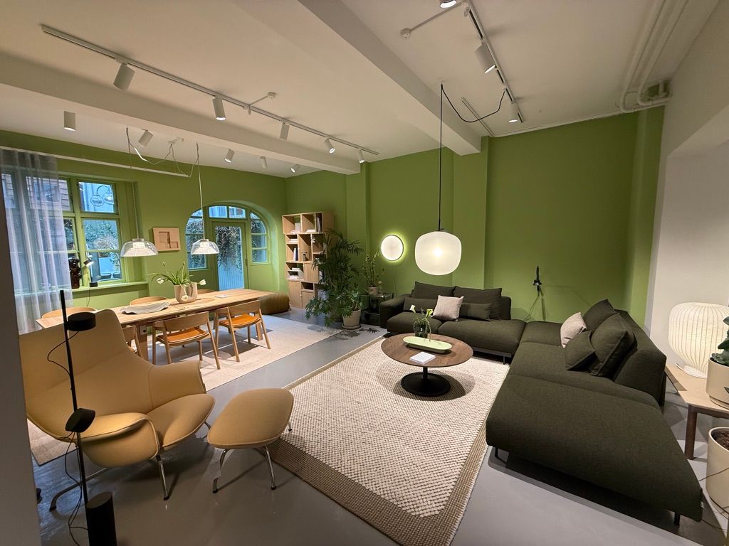 Butik med grønne vægge, møbler og højhængende loftslamper, plante i krukke og bord med blomster.
