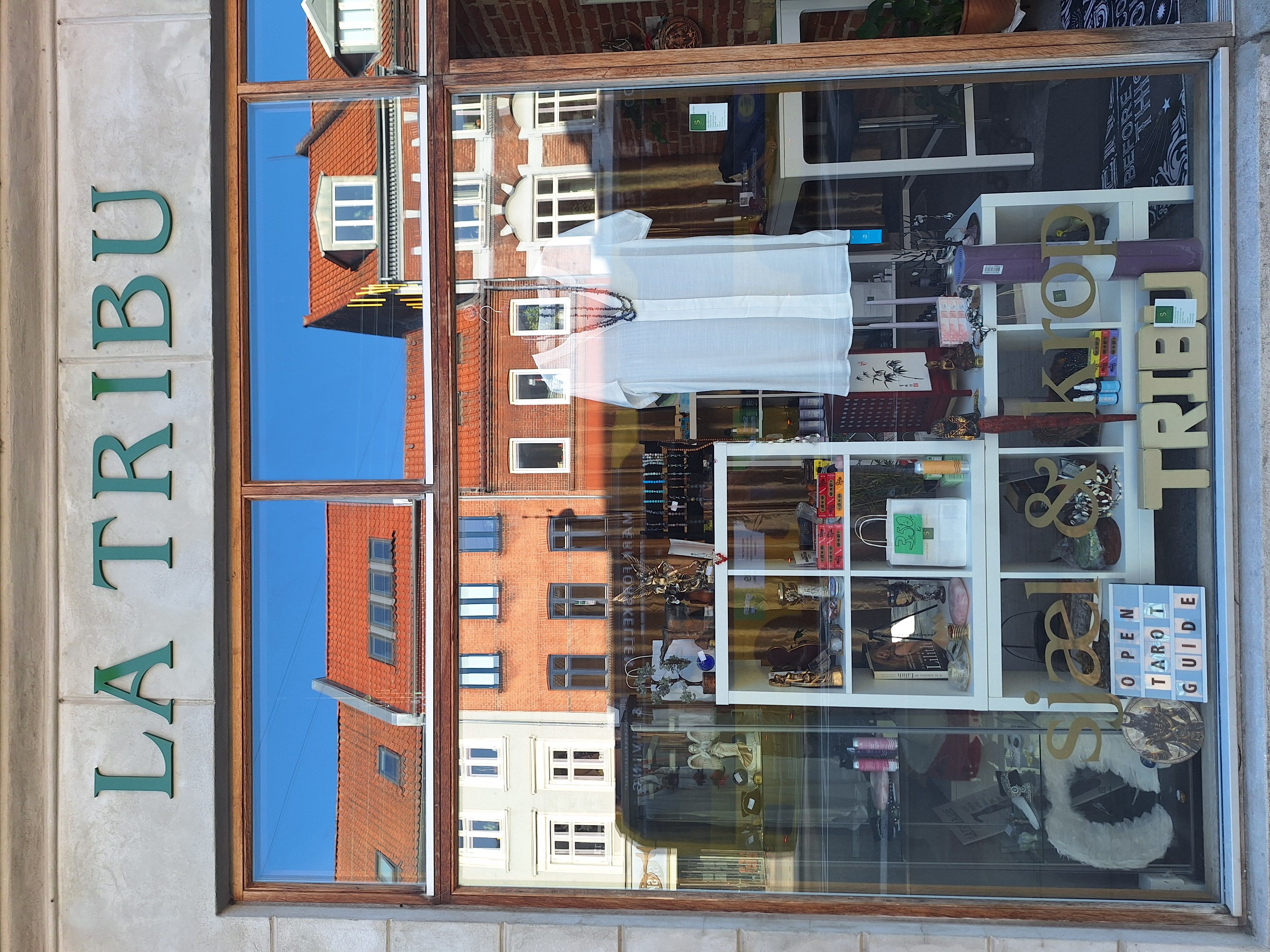 Vindue med refleksioner af bygninger og en indre butiksudsmykning med mærket La Tribu og Reiki Shop.