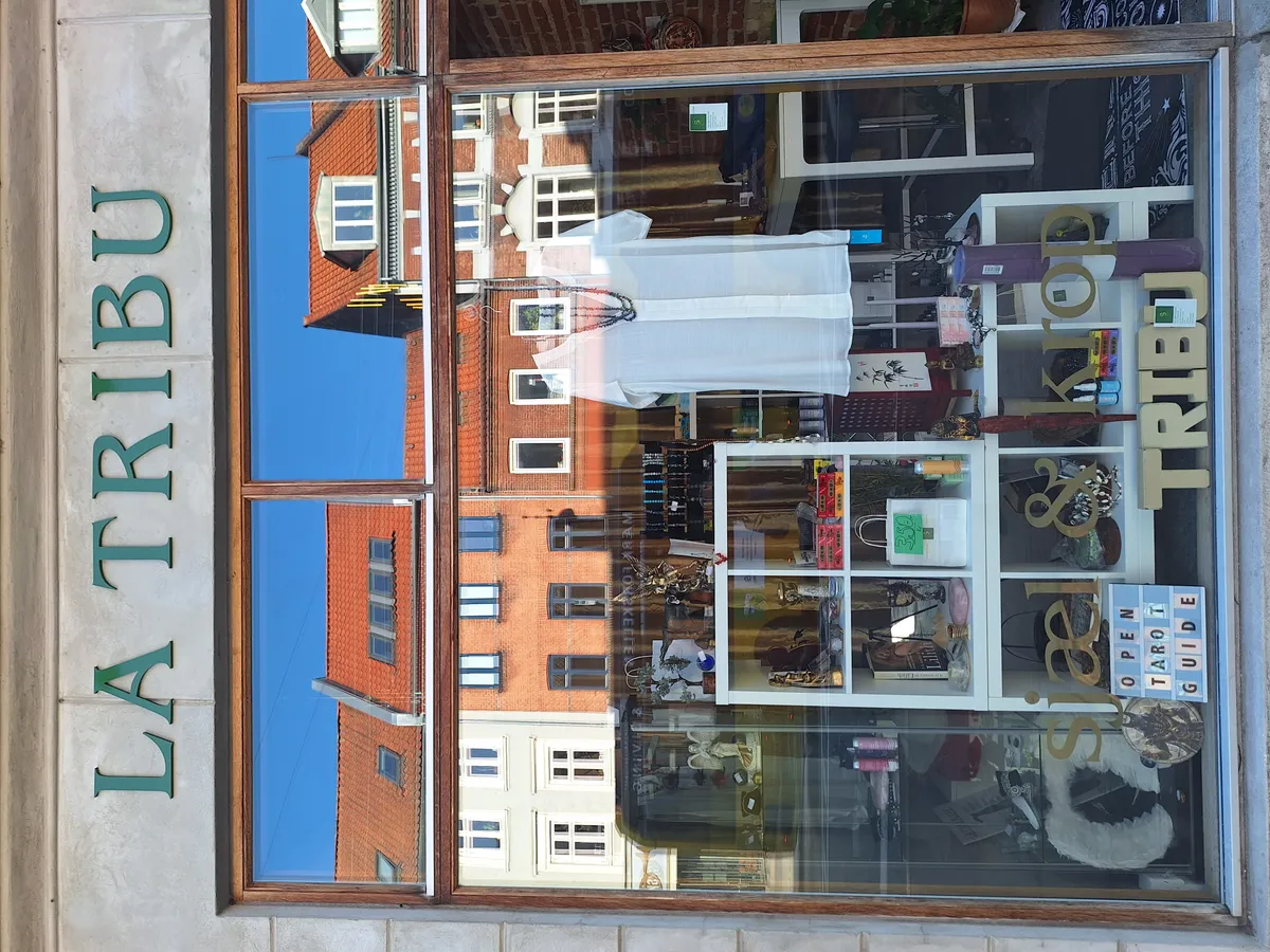 Vindue med refleksioner af bygninger og en indre butiksudsmykning med mærket La Tribu og Reiki Shop.