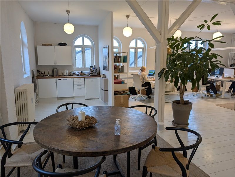 Et moderne kontor med rundt bord, hvide vægge, lysekroner og en stor plante. Der er høj teknologi og to mennesker, der