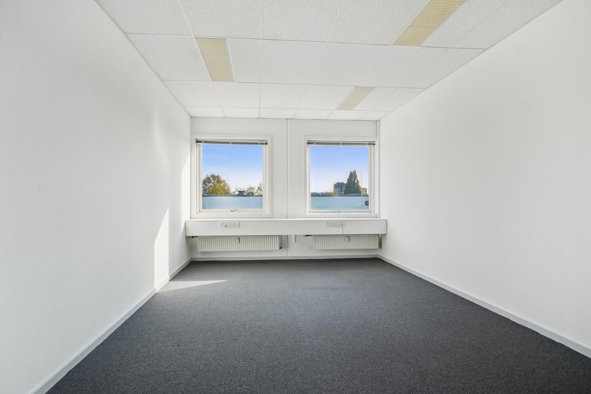 Et tomt kontor med hvide vægge, grå gulvbelægning og to store vinduer med udsigt til bygninger og træer. Der er radiatorer
