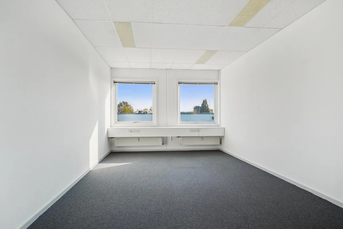 Et tomt kontor med hvide vægge, grå gulvbelægning og to store vinduer med udsigt til bygninger og træer. Der er radiatorer