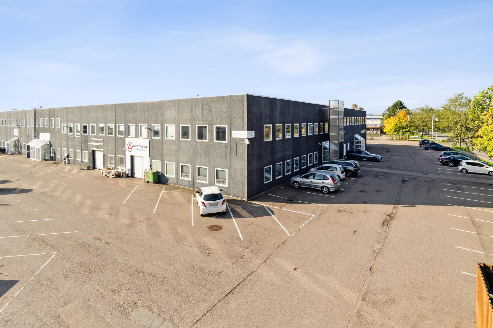 Kontorbyggeri med navneplade ABA Teknik. Foran bygningen står en hvid bil, og der er parkeringspladser med parkerede biler.