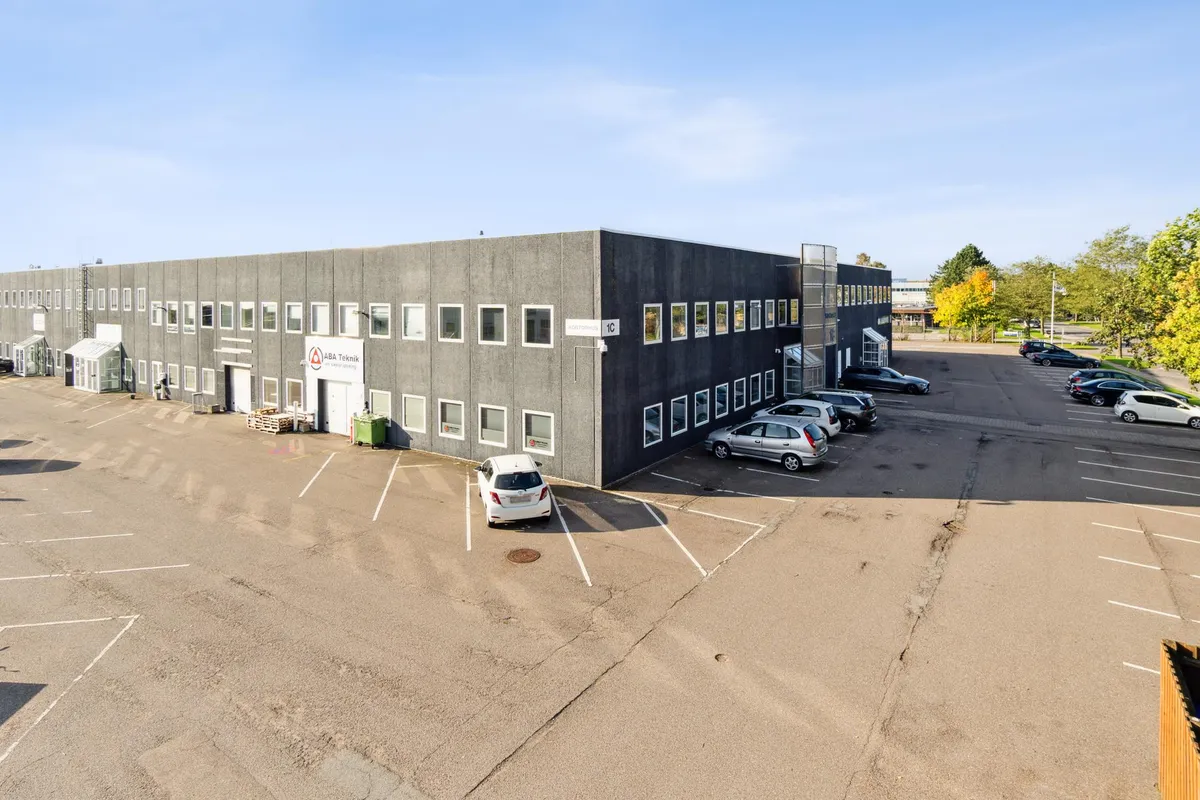 Kontorbyggeri med navneplade ABA Teknik. Foran bygningen står en hvid bil, og der er parkeringspladser med parkerede biler.