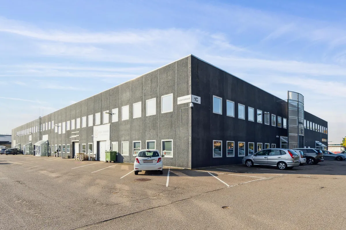 En stor sort bygning med navnet "Kontorhus" og et vindue med en skilt med nummer 1C. Parkerede biler og en grøn container er