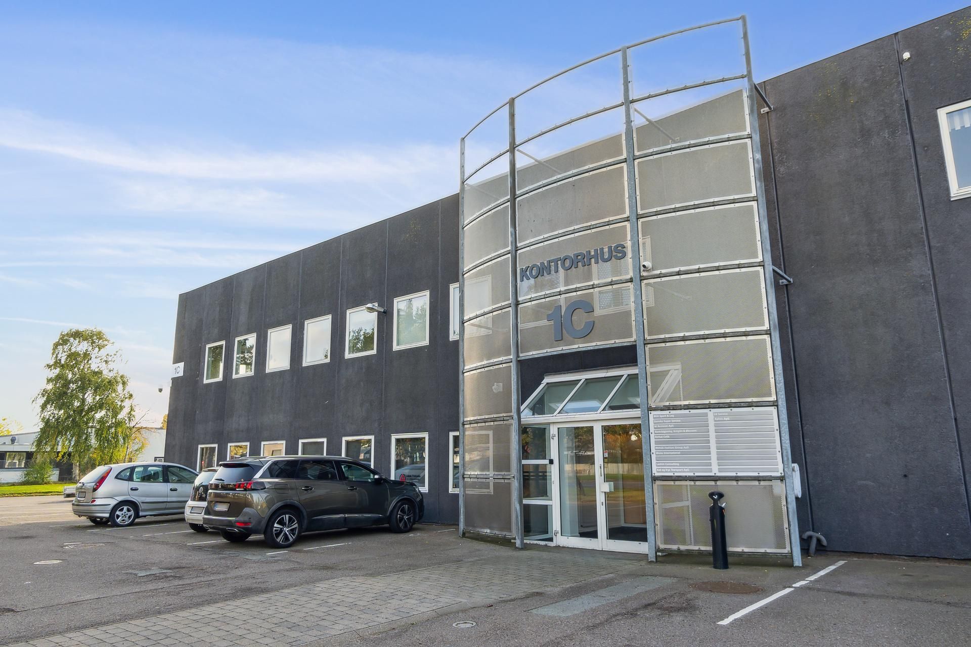 Bygning med glassfasade og teksten Kontorhus 1C, hvor der er parkeret to sorte og en sølv farvet bil foran