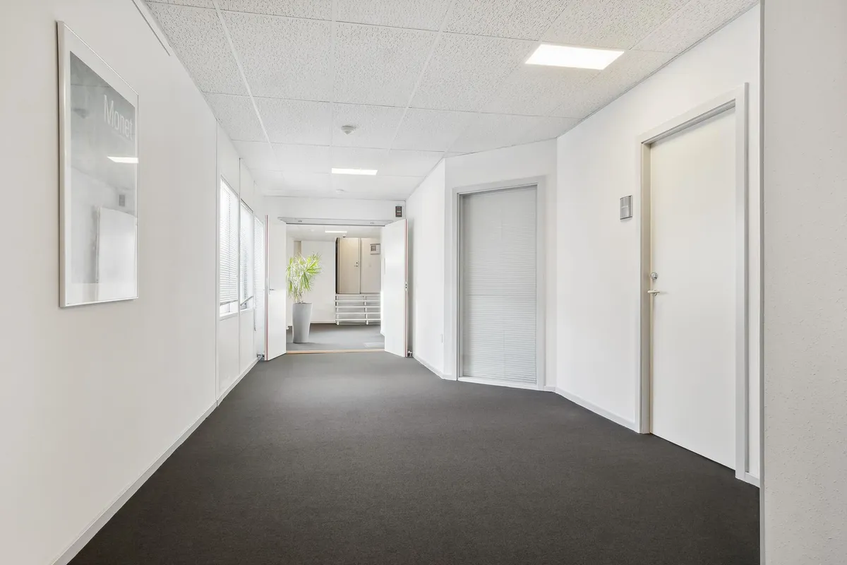 En lang, glasklædt korridor med hvide vægge, hvor det er muligt at se ind i et kontorlokum med hvide vægge, et hvidt loft og