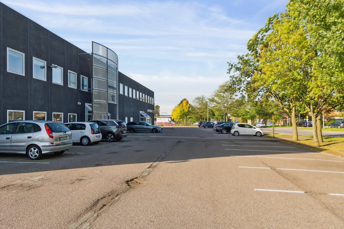 Parkeringsplads med flere parkerede biler, herunder en hvid Nissan og en grå Renault, i forgrunden er en bygning med flere