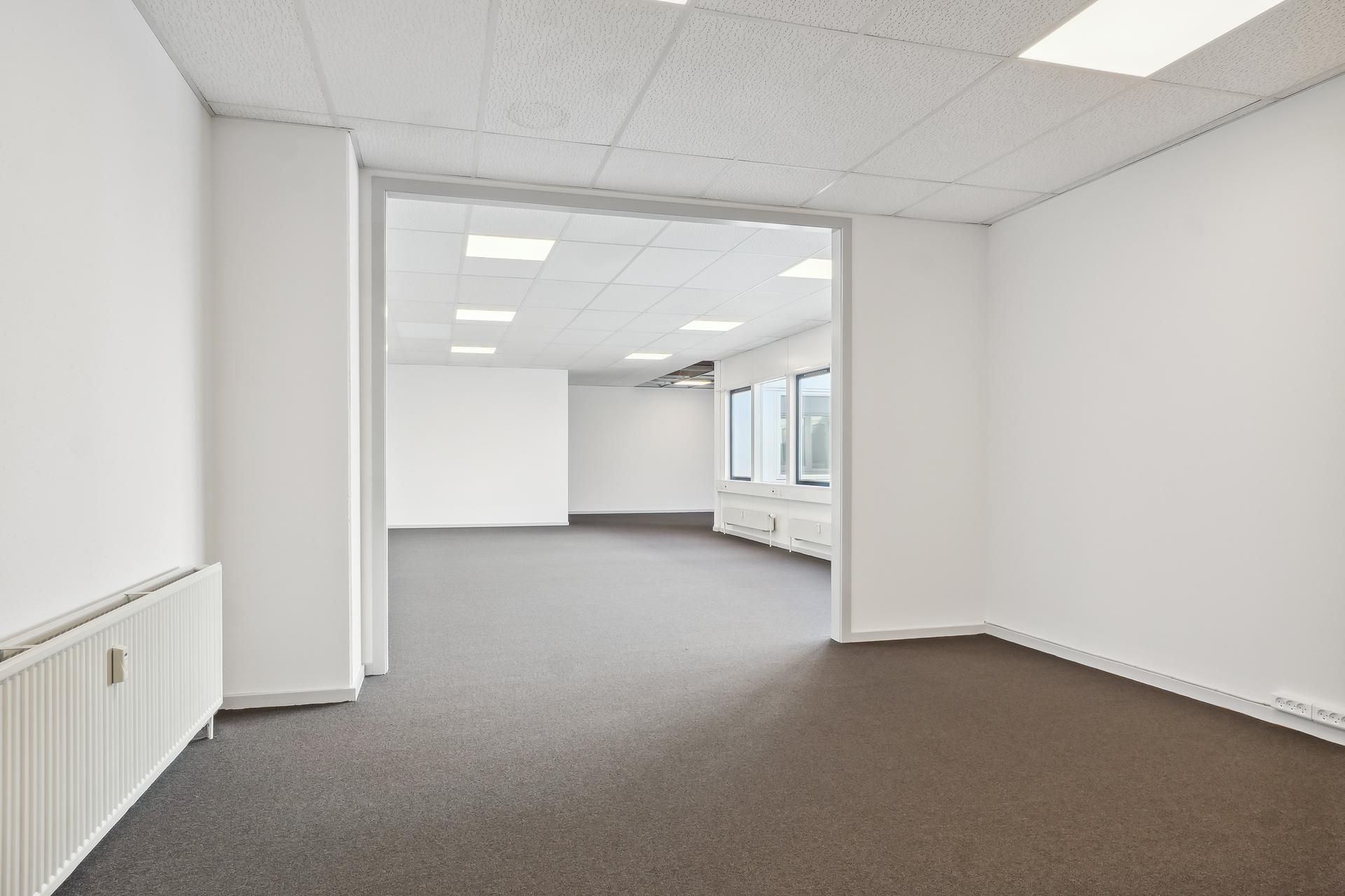 Et tomt kontor med lyse lofter og vægge, gulvtæppe og radiatorer på væggen, hvor vinduer giver masser af naturlys