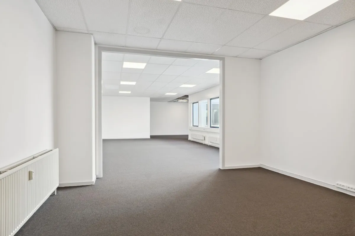 Et tomt kontor med lyse lofter og vægge, gulvtæppe og radiatorer på væggen, hvor vinduer giver masser af naturlys