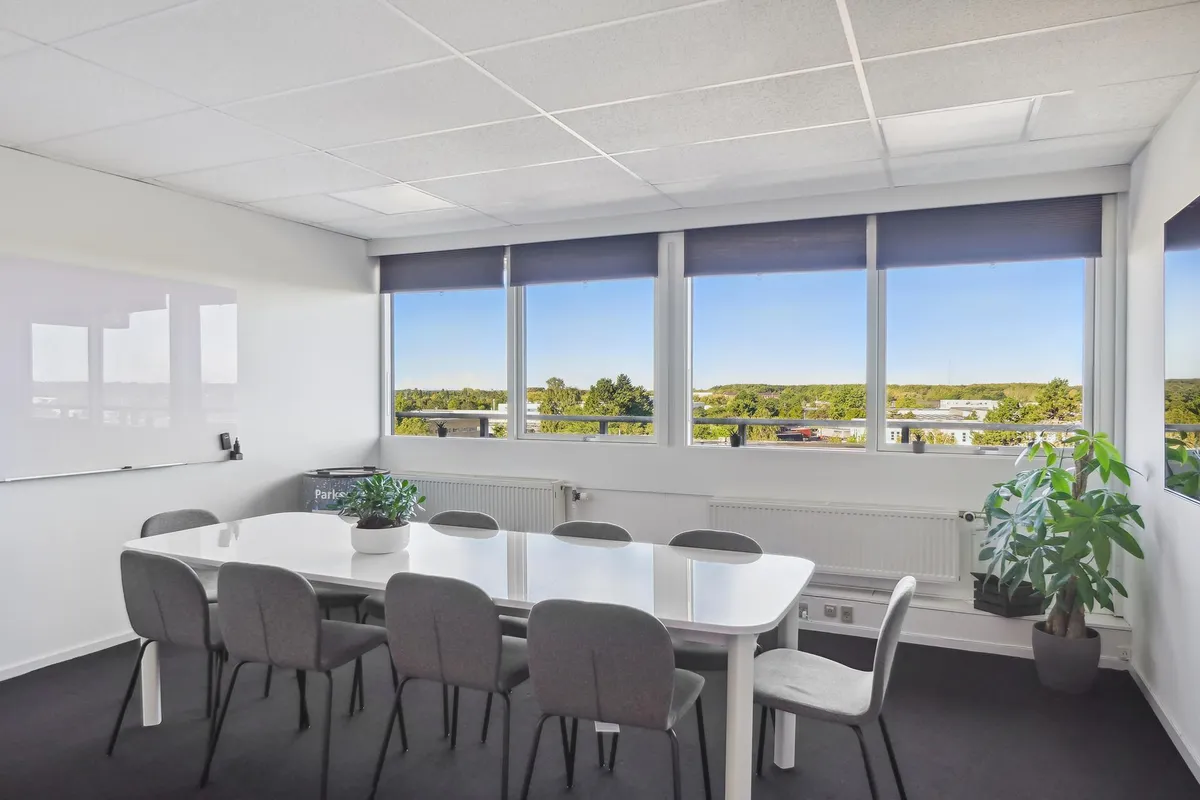 Kontorrum med lyse vinkler og møbler. Et hvidt bord med flere sæder og en plante i hjørnet. To planter og radiatorer langs