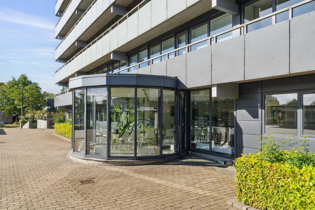 Moderne kontorbygning med glassveranda, der indeholder en plante og møbler, hvor skyerne og træerne i baggrunden tilføjer