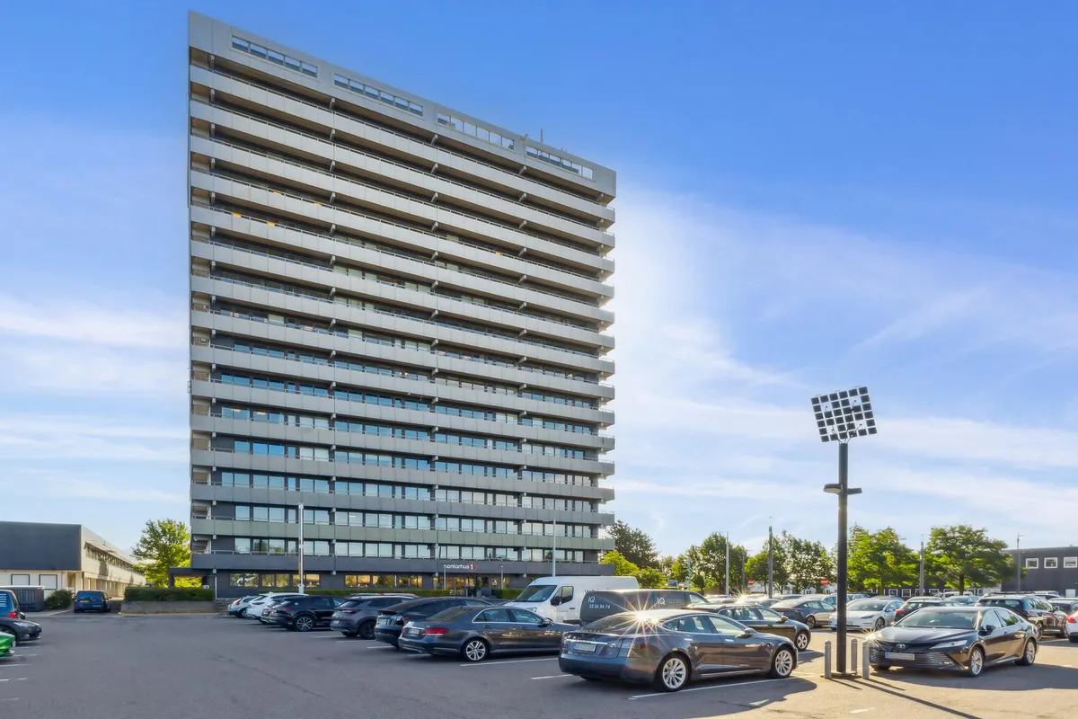 Kontorbyggeri med parkeringsplads og biler parkeret i rækker. Der er en solcelle på en lygte og et skilt med teksten