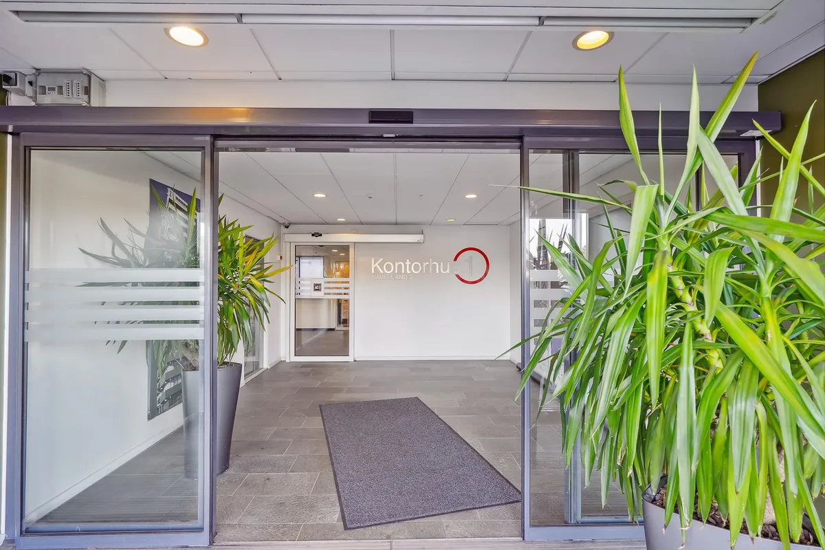 En kontorindgang med to store glasdøre og en gulvbeklædning i brun. Indenfor ses et lobbyområde med en tæppe og to store