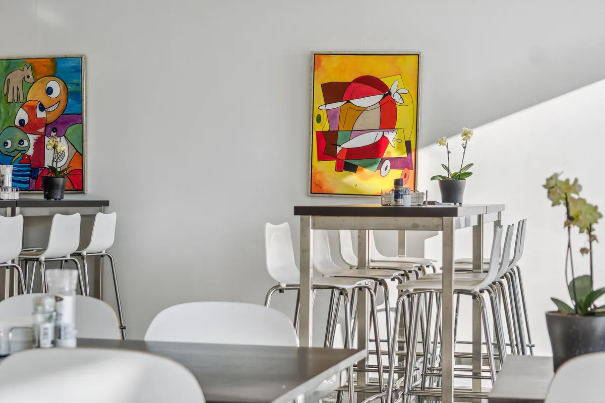 Moderne kontor med hvide stole, bord og to store malerier med farverige abstrakte motiver og plantepotter på væggen