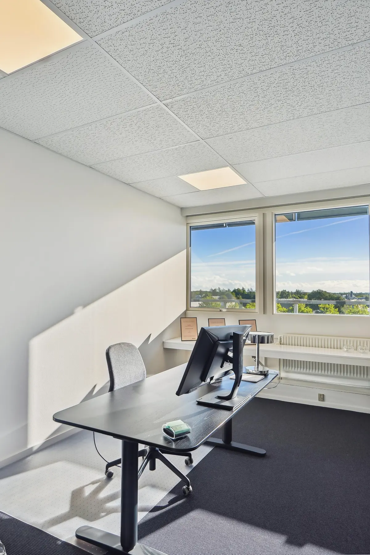 Kontor med store vinduer, to skærme, et bord og en stol og en skab med radiatorer i baggrunden