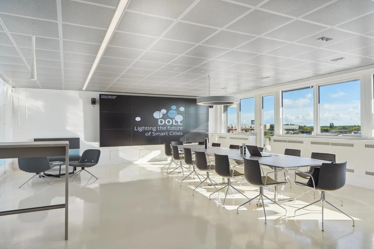 Kontorrum med lyse lofter og store vinduer, møbler og en stor sort skærm med teksten Doll og "Lighting the future of Smart