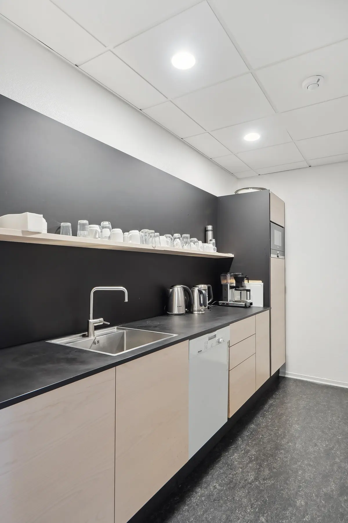 Moderne kontor med lyse lofter, sort væg og møbleret køkken med mikrobølgeovn, kaffemaskine og lyse træskabe.