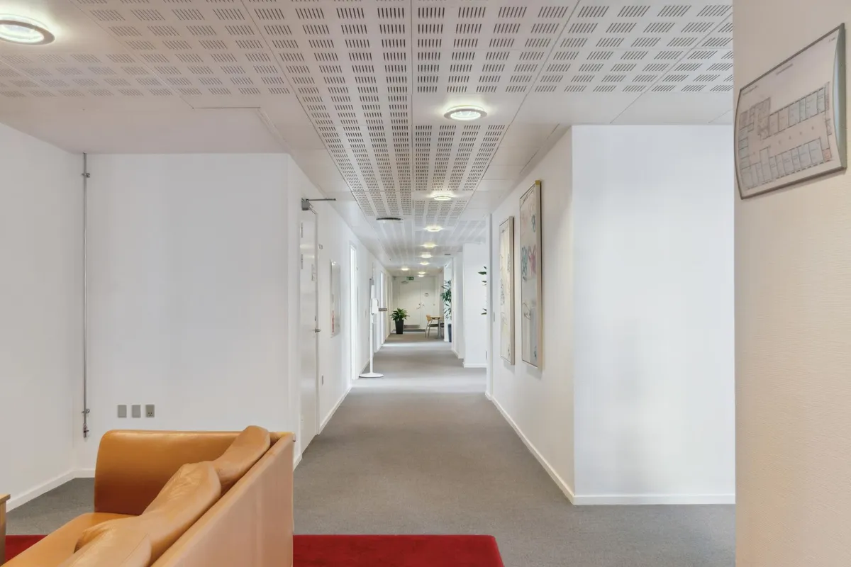 Kort og ren kontorhaller med lyse loftslampe, lysegrå gulv og hvide vægge samt lysebrune lædersofaer og rødt tæppe.
