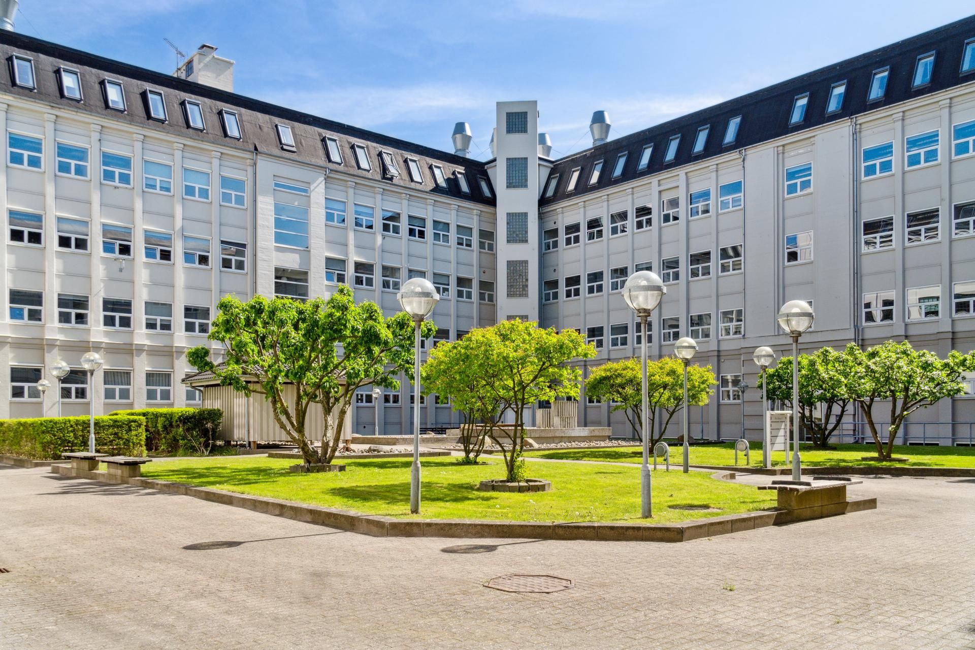 En kontorvæg med høj glasvinduer og gulve, omgivet af blomstrende træer og buske. Foran er en grøn plæne med bænke og