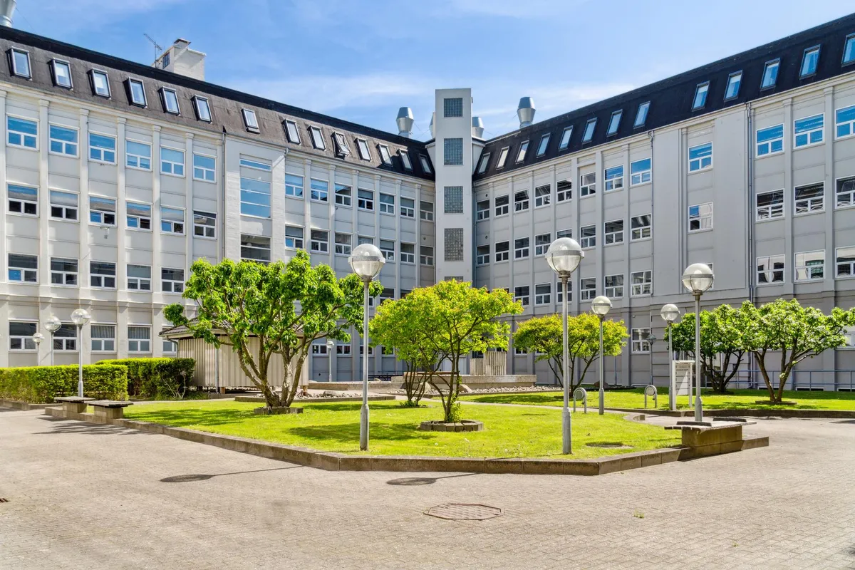 En kontorvæg med høj glasvinduer og gulve, omgivet af blomstrende træer og buske. Foran er en grøn plæne med bænke og