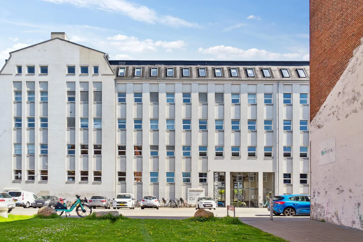 Kontorbygning med fire etager og mange vinduer parkeret foran flere biler og cykler samt en forhøjet græsplæne og en søjle