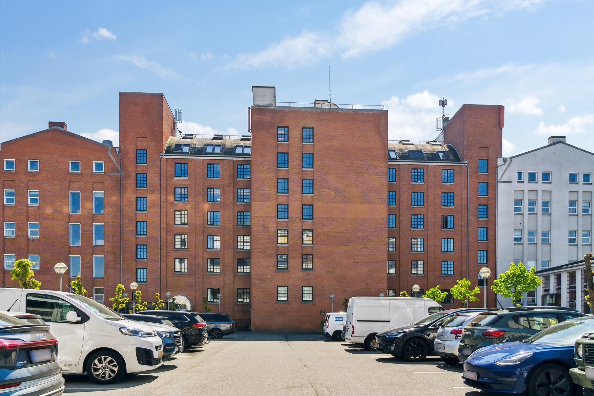 Parkeringsplads foran et stort, rødt bygningskompleks med flere etager og mange vinduer, samt parkerede biler og træer