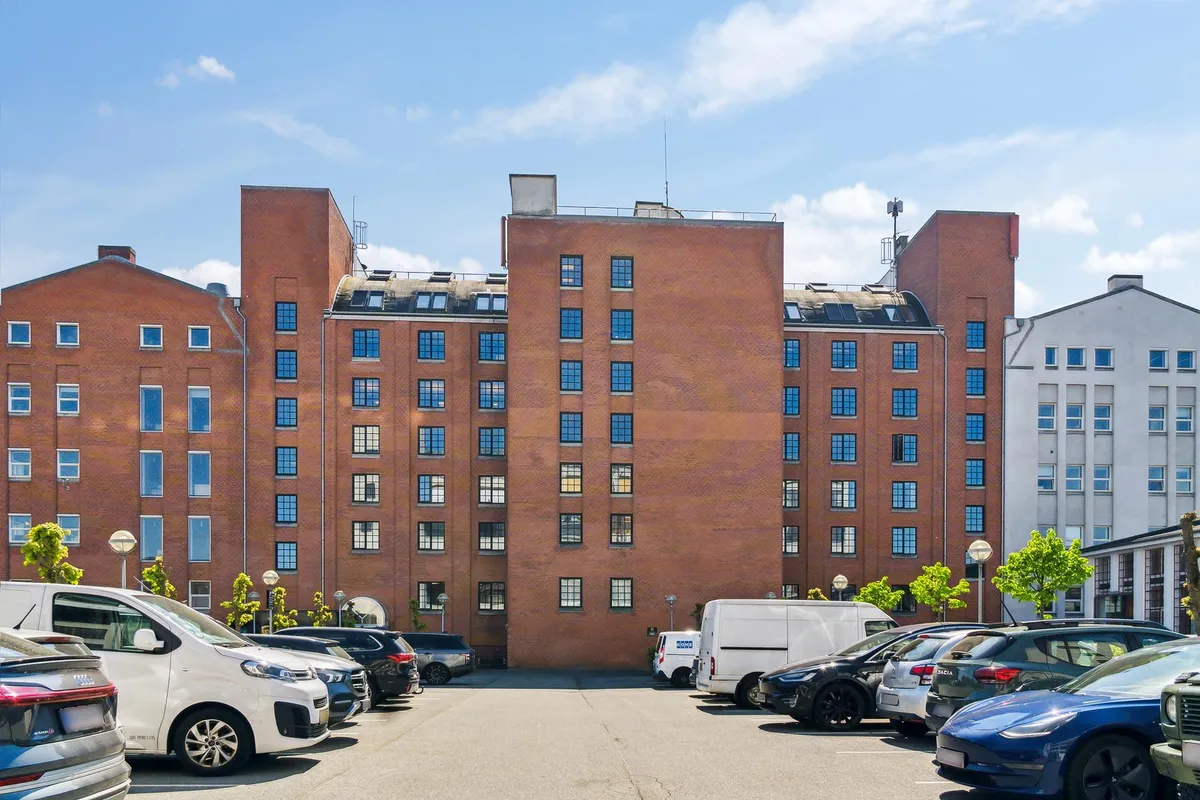 Parkeringsplads foran et stort, rødt bygningskompleks med flere etager og mange vinduer, samt parkerede biler og træer