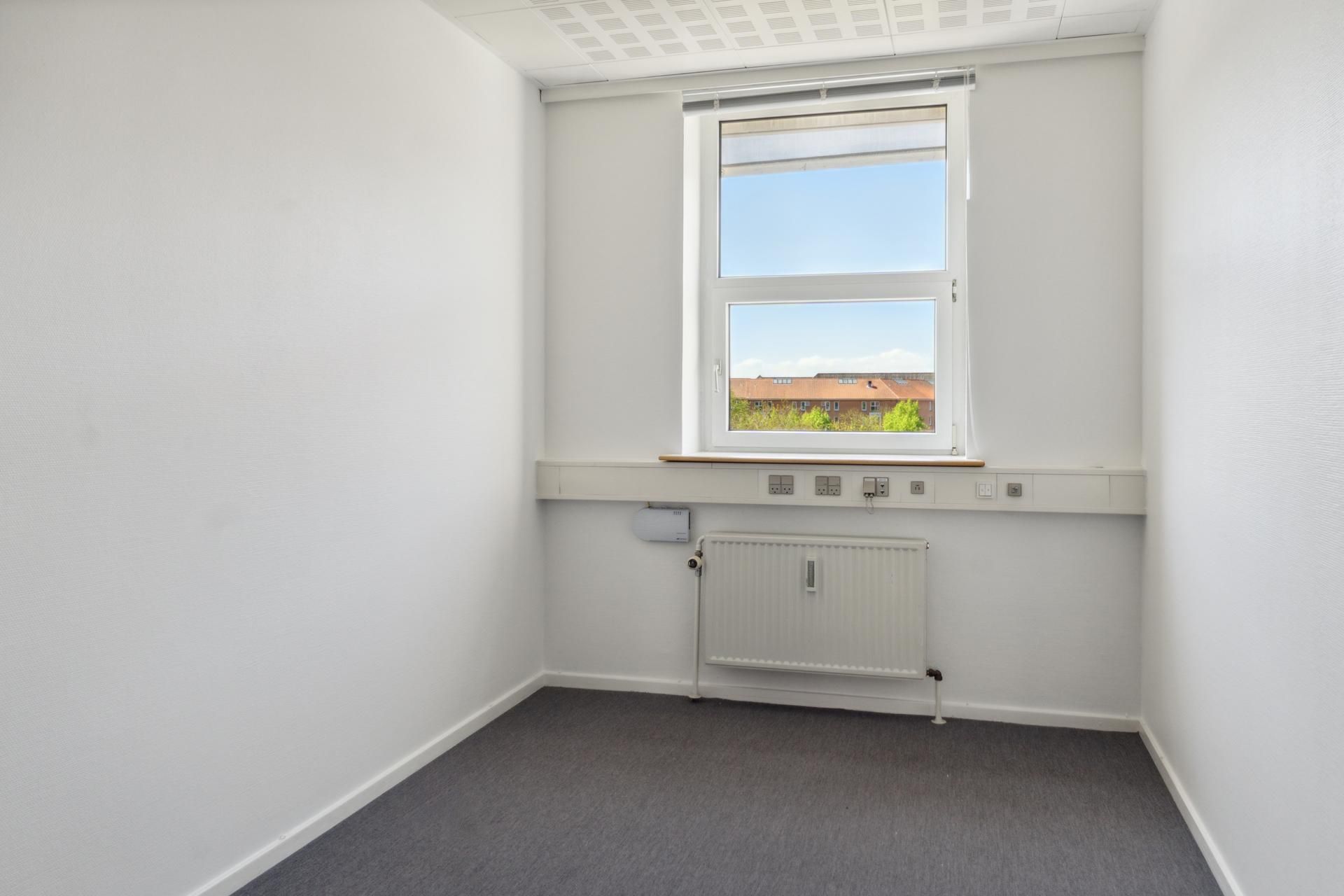 En tom kontorværelse med hvide vægge, hvor vinduet er åbent og viser bygninger og grønne træer i baggrunden